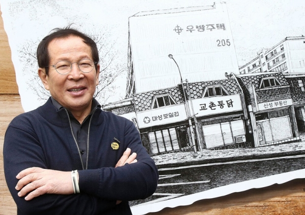 교촌치킨의 창업주인 권원강 교촌에프앤비㈜ 전 회장이 교촌치킨 1호점 일러스트 앞에서 사진 촬영을 하고 있는 모습. 사진=교촌.
